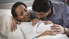 A person laying in a hospital bed with a newborn on their lap and another person standing beside them
