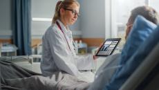 Healthcare provider sitting on a patient's hospital bed while holding a tablet showing diagnostic images of lungs