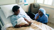 Healthcare provider sitting next to a hospital bed with a teenager in it looking at a phone